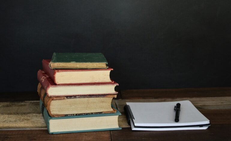 Stack of vintage books next to a modern notebook and pen against a dark background.
